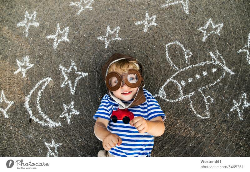 Porträt eines lächelnden Kleinkindes mit Pilotenhut und Schutzbrille, das auf Asphalt liegt und mit Flugzeug, Mond und Sternen bemalt ist Flieger Flugzeuge