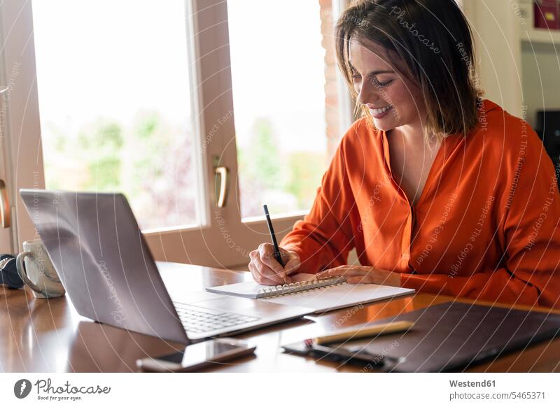 Lächelnde Geschäftsfrau schreibt bei der Arbeit zu Hause auf den Notizblock Farbaufnahme Farbe Farbfoto Farbphoto Innenaufnahme Innenaufnahmen innen drinnen Tag
