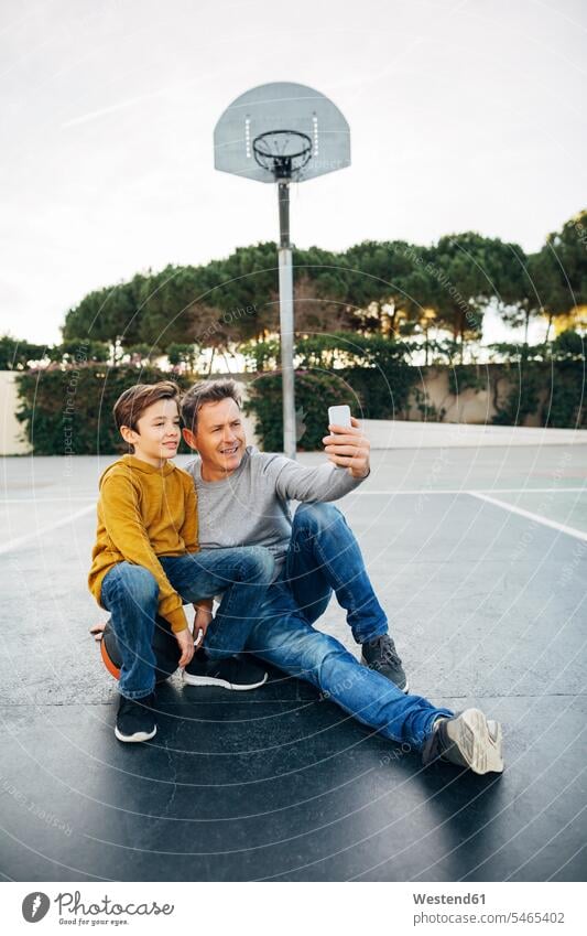 Vater und Sohn sitzen auf einem Basketballplatz im Freien und machen ein Selfie Sportplatz Sportplaetze Sportplätze sitzend sitzt Papas Väter Vati Vatis Papis
