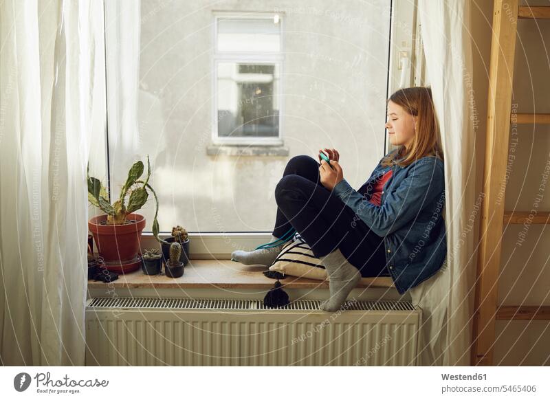 Mädchen sitzt zu Hause mit Smartphone am Fenster Blumentoepfe Blumentöpfe Telekommunikation telefonieren Handies Handys Mobiltelefon Mobiltelefone Lektüre