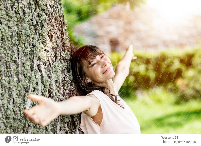Porträt einer lächelnden brünetten Frau, die sich gegen einen Baumstamm lehnt Leute Menschen People Person Personen Europäisch Kaukasier kaukasisch 1 Ein