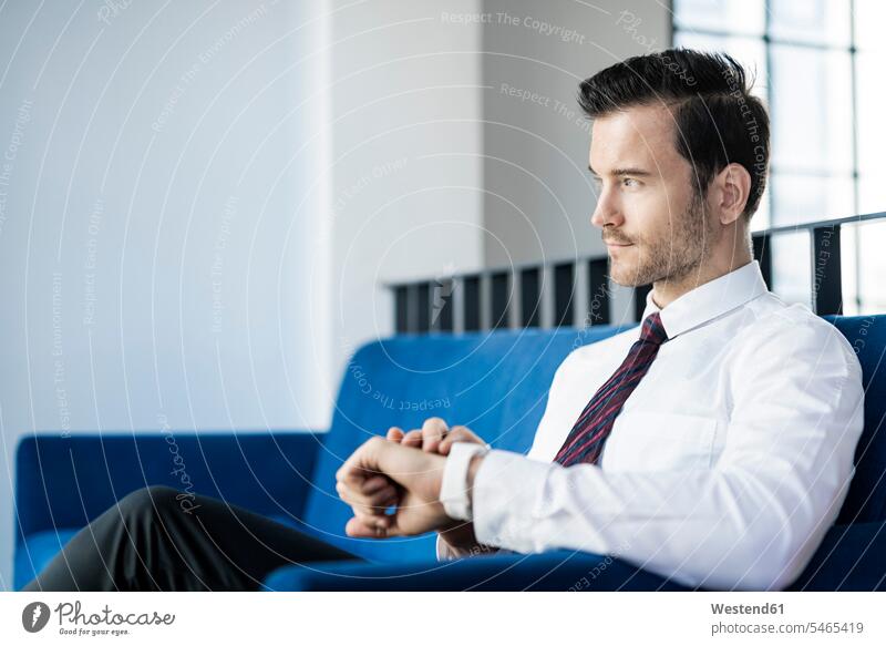 Geschäftsmann sitzt auf blauer Couch im Büro und überprüft seine Smartwatch checken sitzen sitzend Businessmann Businessmänner Geschäftsmänner Office Büros