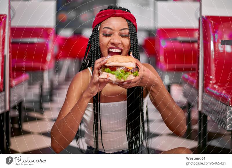 Junge Frau mit geflochtener Frisur sitzt auf dem Boden und greift mit beiden Händen nach einem großen Hamburger sitzend auf dem Boden sitzend gross Grund Land