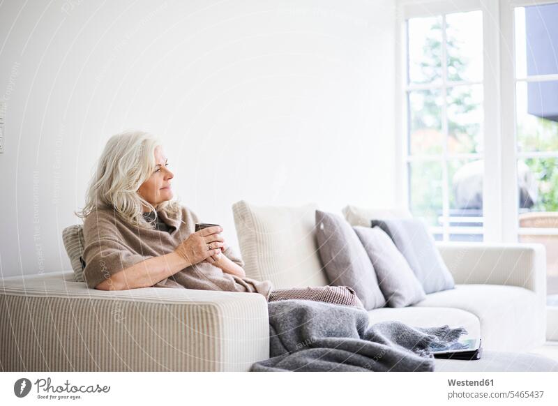 Nachdenkliche Frau mit langen weißen Haaren hält Kaffeetasse beim Sitzen auf dem Sofa in der Wohnung Farbaufnahme Farbe Farbfoto Farbphoto Dänemark Skandinavien