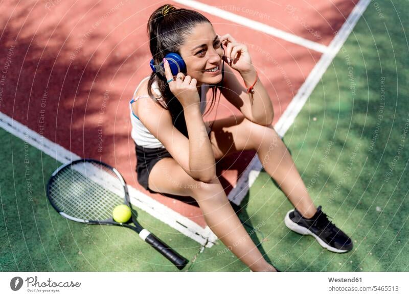Lächelnde Tennisspielerin hört Musik, während sie auf dem Boden des Courts sitzt Farbaufnahme Farbe Farbfoto Farbphoto Spanien Außenaufnahme außen draußen