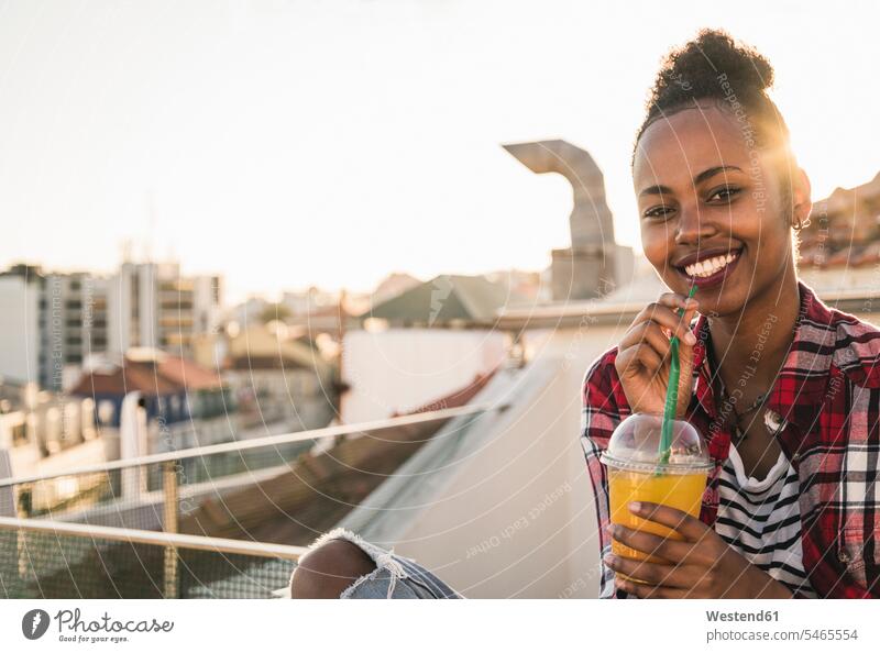 Porträt einer glücklichen jungen Frau, die bei Sonnenuntergang auf dem Dach einen Drink nimmt Leute Menschen People Person Personen Afrikanisch