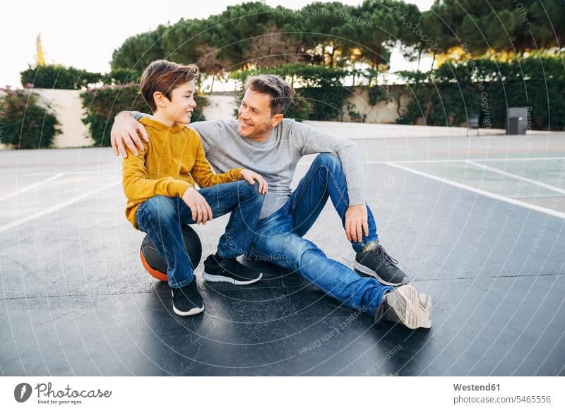 Vater umarmt Sohn auf Basketballfeld Sportplatz Sportplaetze Sportplätze Basketbaelle Basketbälle sitzen sitzend sitzt Söhne umarmen Umarmung Umarmungen