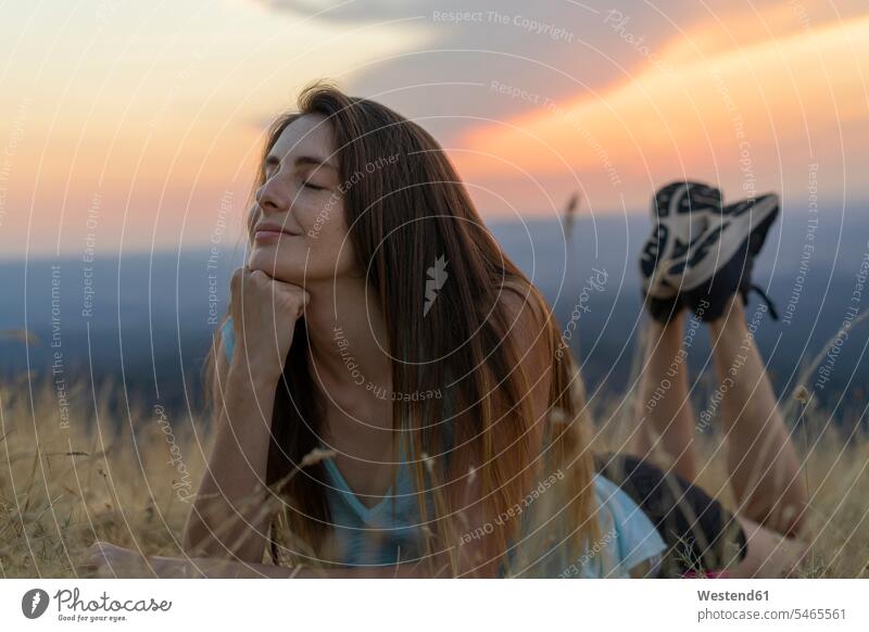Lächelnde junge Frau mit geschlossenen Augen bei Sonnenuntergang im Gras liegend liegt lächeln Gräser Graeser weiblich Frauen Sonnenuntergänge Erwachsener