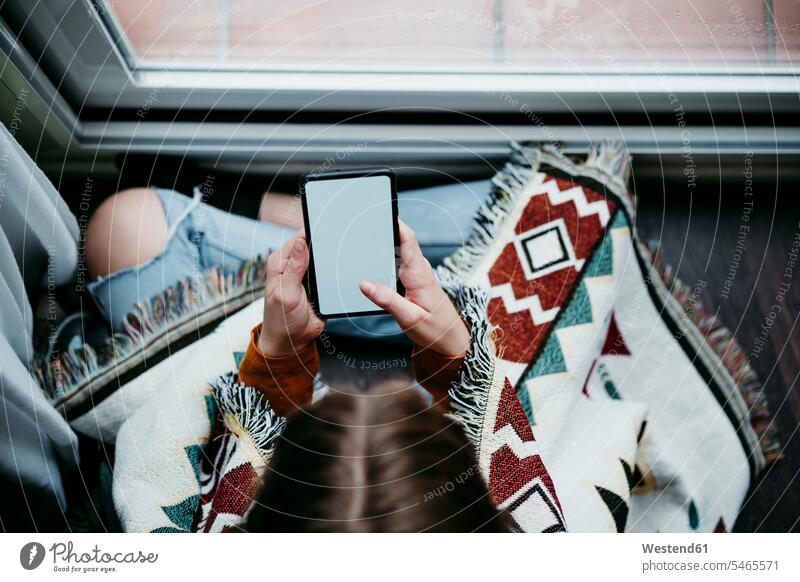 Mädchen mit Decke benutzt Mobiltelefon, während sie zu Hause am Fenster sitzt Farbaufnahme Farbe Farbfoto Farbphoto Innenaufnahme Innenaufnahmen innen drinnen