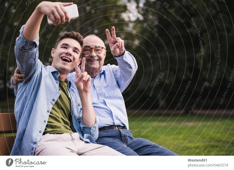 Älterer Mann und Enkel sitzen zusammen auf einer Parkbank und machen ein Selfie mit Smartphone Leute Menschen People Person Personen Europäisch Kaukasier