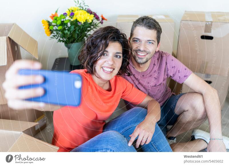 Glückliches Paar macht ein Selfie im neuen Haus Kartons Pappkarton Pappkartons T-Shirts Telekommunikation telefonieren Handies Handys Mobiltelefon Mobiltelefone