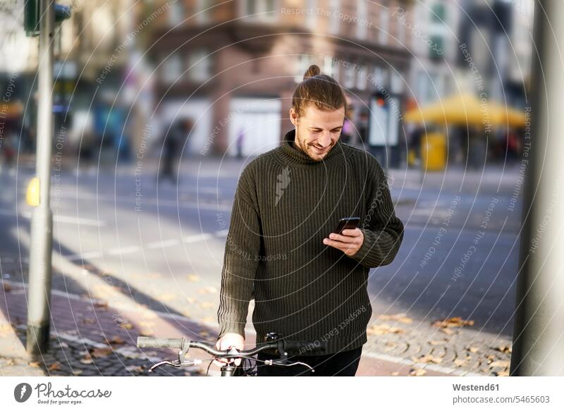 Lächelnder junger Mann mit Fahrrad in der Stadt schaut auf Handy ansehen Männer männlich schauen sehend lächeln Smartphone iPhone Smartphones Bikes Fahrräder