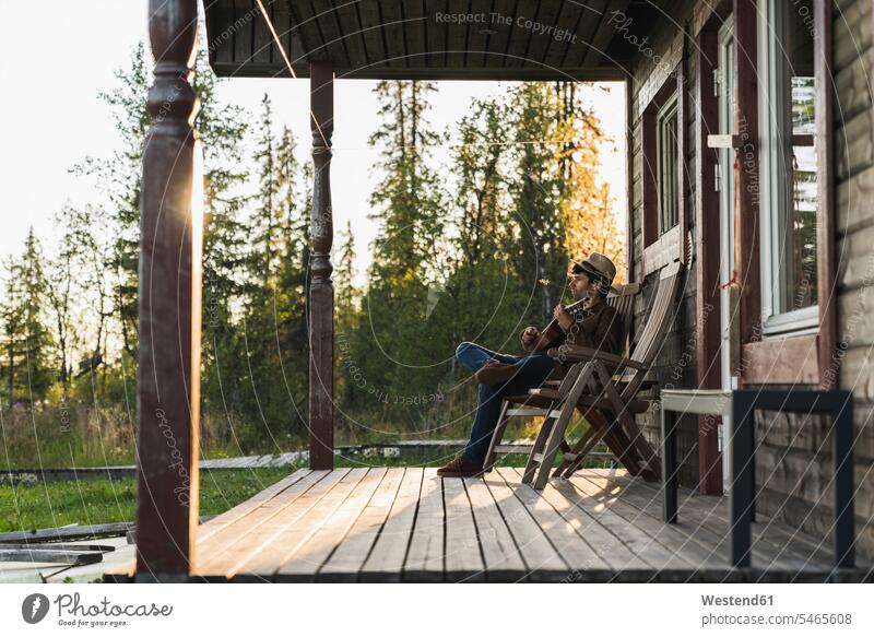 Junger Mann sitzt auf der Veranda eines Holzhauses und spielt Ukulele Patio Vorbau spielen sitzen sitzend Holzhäuser Holzhaeuser romantisch schwärmerisch