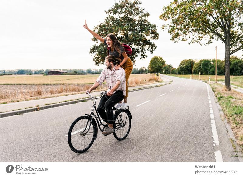 Glückliches junges Paar fährt gemeinsam auf einem Fahrrad auf der Landstraße Bikes Fahrräder Räder Rad Landstraßen fahren Gemeinsam Zusammen Miteinander Pärchen