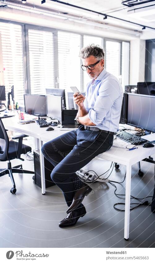 Reifer Geschäftsmann sitzt im Büro am Schreibtisch und benutzt ein Smartphone sitzen sitzend Office Büros iPhone Smartphones Arbeitstisch Schreibtische