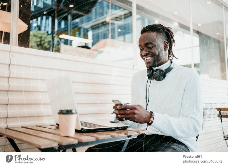 Lächelnde kreative Geschäftsmann mit Laptop mit Handy im Café Farbaufnahme Farbe Farbfoto Farbphoto Außenaufnahme außen draußen im Freien Tag Tageslichtaufnahme