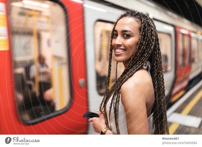 Porträt einer glücklichen jungen Frau, die am Bahnsteig einer U-Bahn-Station wartet, London, Großbritannien Leute Menschen People Person Personen gemischtrassig