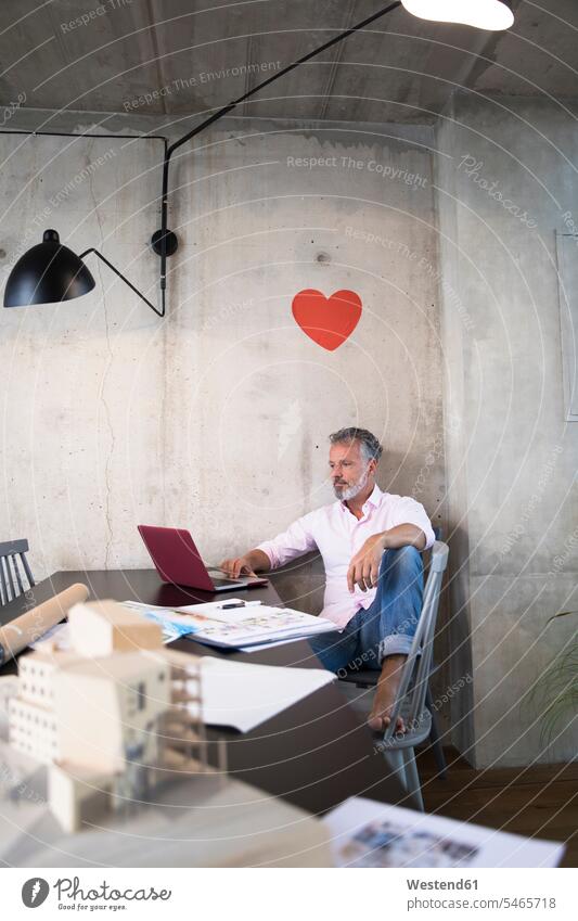Geschäftsmann in einem Loft mit Laptop mit Dokumenten und Architekturmodell auf dem Tisch und Herz an der Wand Lofts Herzform Herzformen Herzen Unterlagen