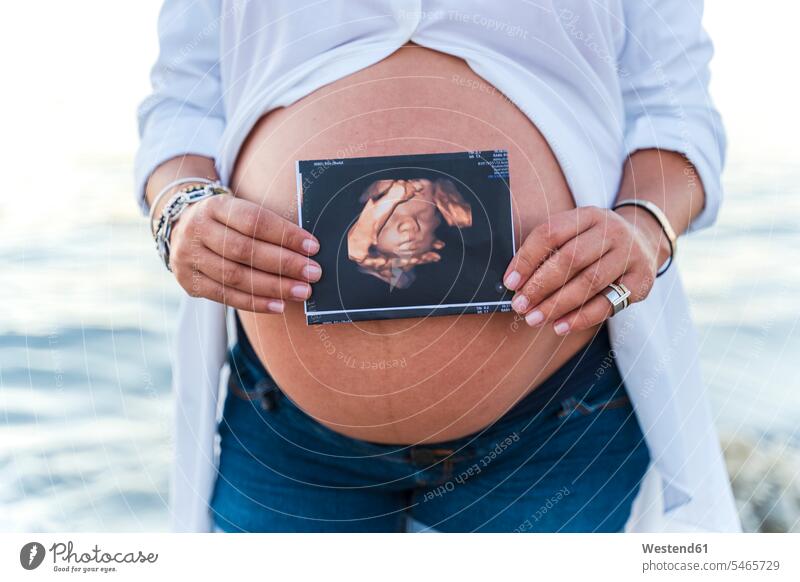 Mittelsektion einer schwangeren Frau, die ein Sonogramm zeigt, während sie vor dem Wasser steht Farbaufnahme Farbe Farbfoto Farbphoto Außenaufnahme außen
