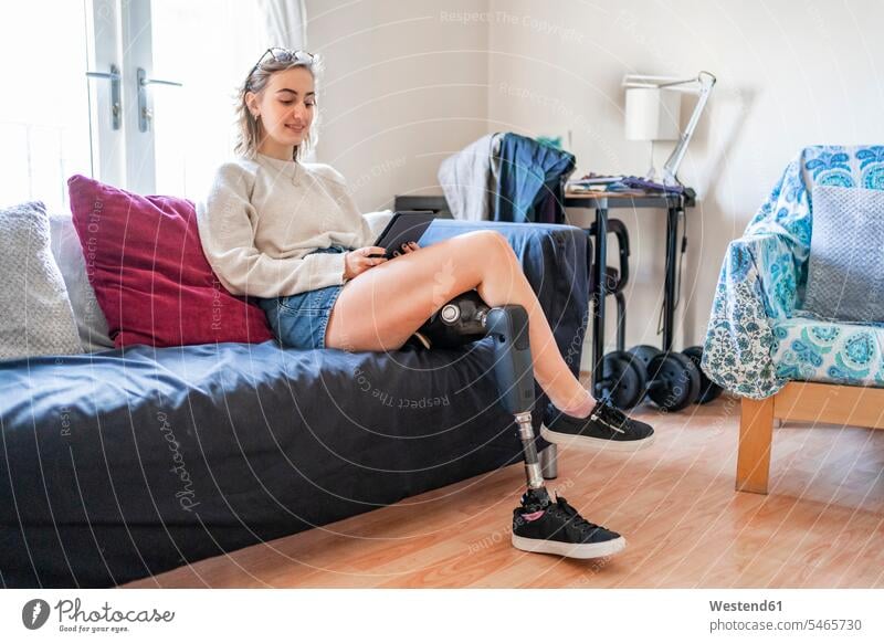 Junge Frau mit Beinprothese sitzt zu Hause auf Couch und liest E-Book Leute Menschen People Person Personen Alleinstehende Alleinstehender Singles