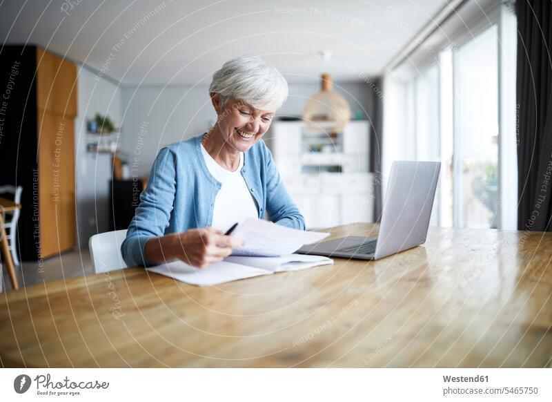 Lächelnde aktive ältere Frau, die zu Hause sitzt und Papierkram erledigt Farbaufnahme Farbe Farbfoto Farbphoto Innenaufnahme Innenaufnahmen innen drinnen Tag
