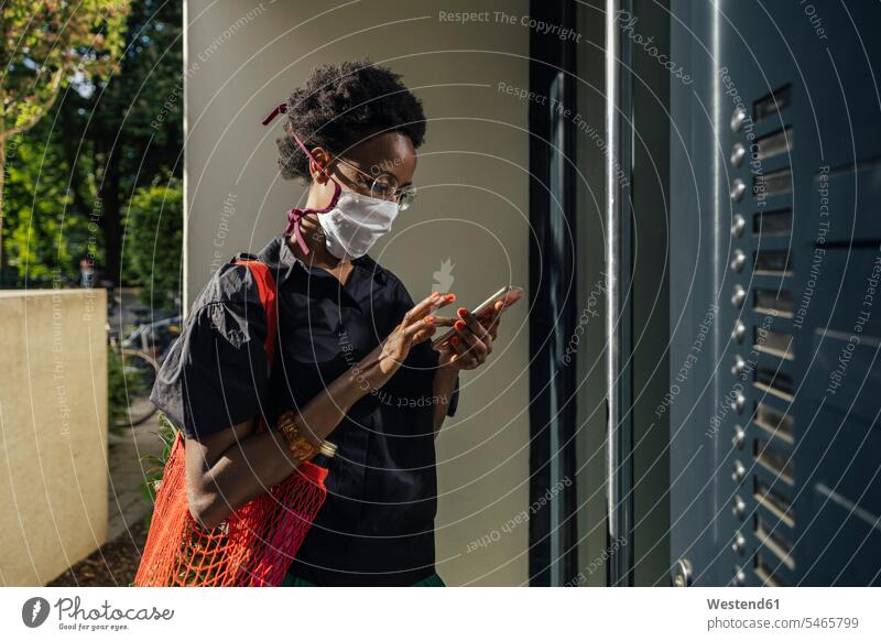 Junge Frau mit textiler Schutzmaske steht mit einem Smartphone vor der Eingangstür Taschen Einkaufstaschen Telekommunikation Handies Handys Mobiltelefon