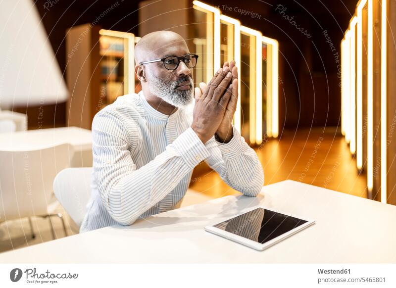 Porträt eines nachdenklichen reifen Mannes, der am Schreibtisch in einer Bibliothek mit digitalem Tablet sitzt Arbeitstisch Schreibtische sitzen sitzend