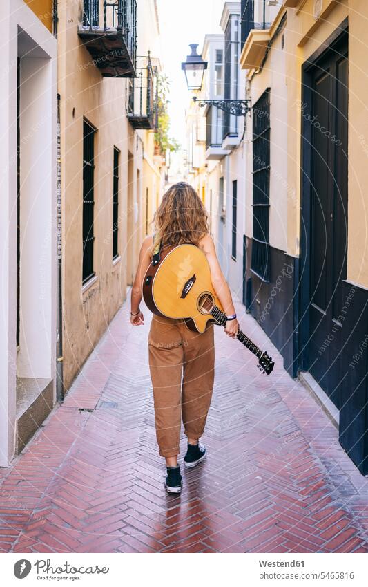 Rückansicht einer jungen Frau mit Gitarre, die auf einer schmalen Straße inmitten von Gebäuden in Santa Cruz, Sevilla, Spanien, spazieren geht Farbaufnahme