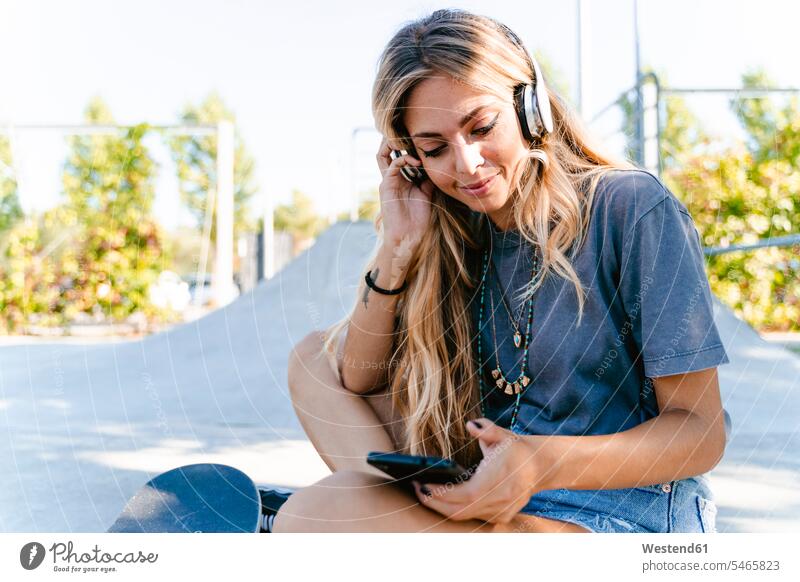 Junge blonde Frau hört Musik über Kopfhörer, während sie im Skateboard-Park ein Smartphone benutzt Farbaufnahme Farbe Farbfoto Farbphoto Außenaufnahme außen