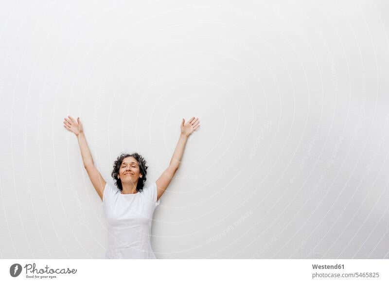 Lächelnde reife Frau steht mit geschlossenen Augen und erhobenen Armen gegen die weiße Wand Farbaufnahme Farbe Farbfoto Farbphoto Spanien Außenaufnahme außen