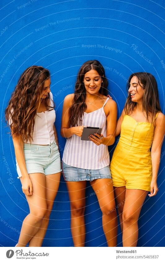 Drei glückliche Freundinnen stehen an einer blauen Wand und teilen sich ein Tablett Freunde Kameradschaft dunkle Haare braunes Haar braunhaarig brünett Medium