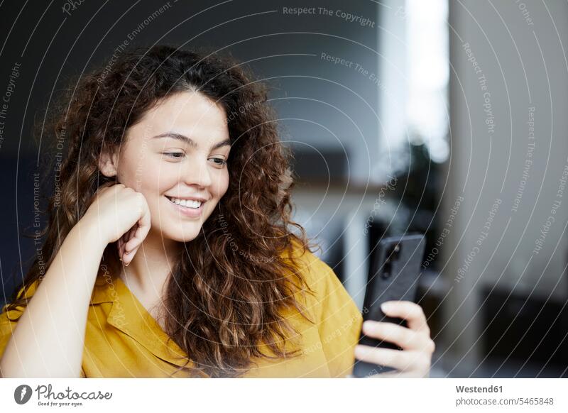 Lächelnde schöne Frau benutzt Smartphone, während sie zu Hause sitzt Farbaufnahme Farbe Farbfoto Farbphoto Innenaufnahme Innenaufnahmen innen drinnen Tag