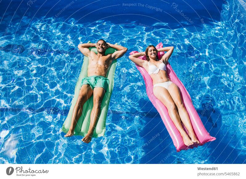 Lächelndes junges Paar mit Händen hinter dem Kopf entspannt auf Luftmatratzen im Schwimmbad Farbaufnahme Farbe Farbfoto Farbphoto Spanien Tag Tageslichtaufnahme
