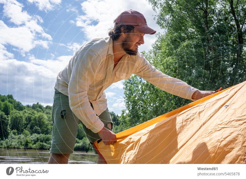 Mittelgroßer erwachsener Mann installiert Zelt im Wald gegen den Himmel auf dem Campingplatz Farbaufnahme Farbe Farbfoto Farbphoto Freizeitbeschäftigung Muße