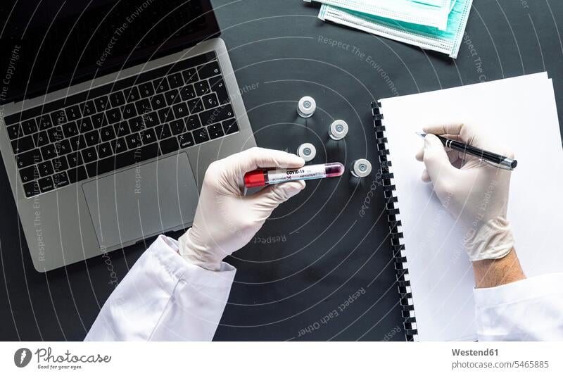 Arzt mit COVID-19-Probenschrift auf Spiralnotebook per Laptop am Schreibtisch im Labor Farbaufnahme Farbe Farbfoto Farbphoto Innenaufnahme Innenaufnahmen innen