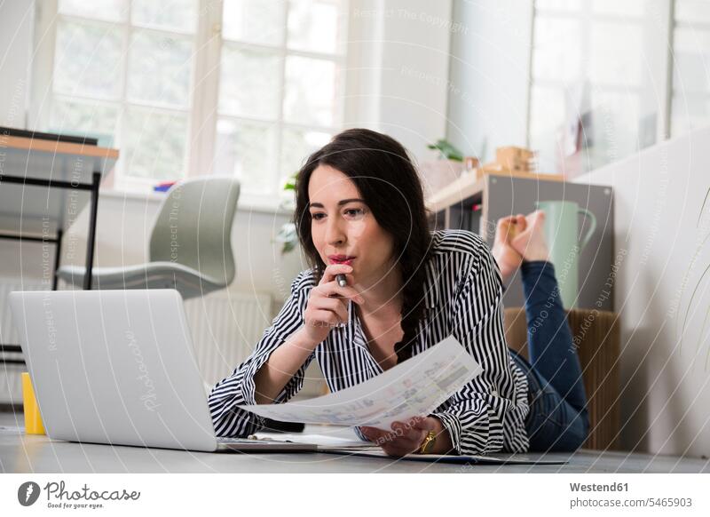 Lässige Frau mit Blatt Papier und Laptop, die im Büro auf dem Boden liegt weiblich Frauen Office Büros liegen liegend Böden Boeden Blaetter Papierblätter