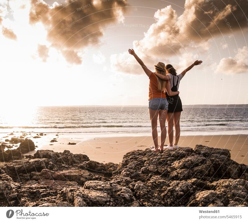 Zwei Freundinnen stehen am felsigen Strand, winken bei Sonnenuntergang, Rückansicht freuen Frohsinn Fröhlichkeit Heiterkeit stehend steht frei Muße Miteinander