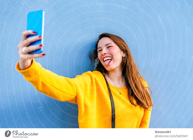 Glückliche junge Frau macht ein Selfie an einer Wand und streckt die Zunge heraus weiblich Frauen Selfies Zungen Mauer Mauern Erwachsener erwachsen Mensch