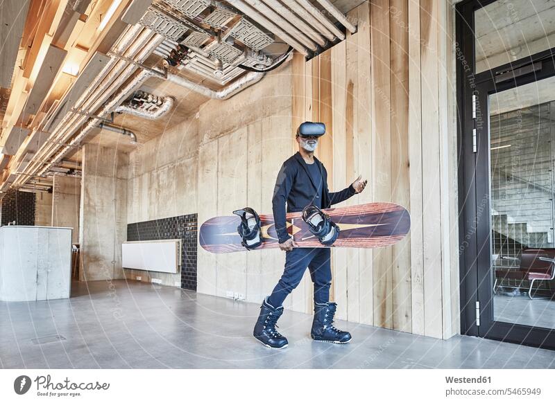 Reifer Geschäftsmann mit VR-Brille hält Snowboard in modernem Büro Businessmann Businessmänner Geschäftsmänner Office Büros virtuell Virtualität Snowboards