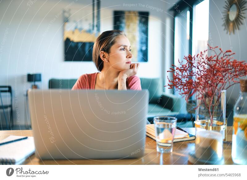 Nachdenkliche junge Frau mit Laptop auf dem Schreibtisch, die weg schaut, während sie zu Hause sitzt Farbaufnahme Farbe Farbfoto Farbphoto Innenaufnahme