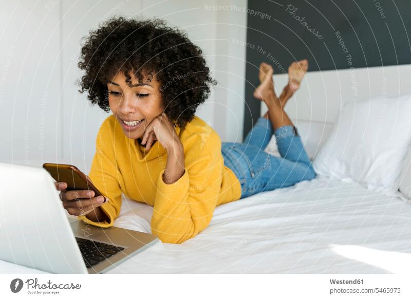 Lächelnde Frau auf dem Bett liegend mit Handy und Laptop Notebook Laptops Notebooks Mobiltelefon Handies Handys Mobiltelefone liegt lächeln weiblich Frauen