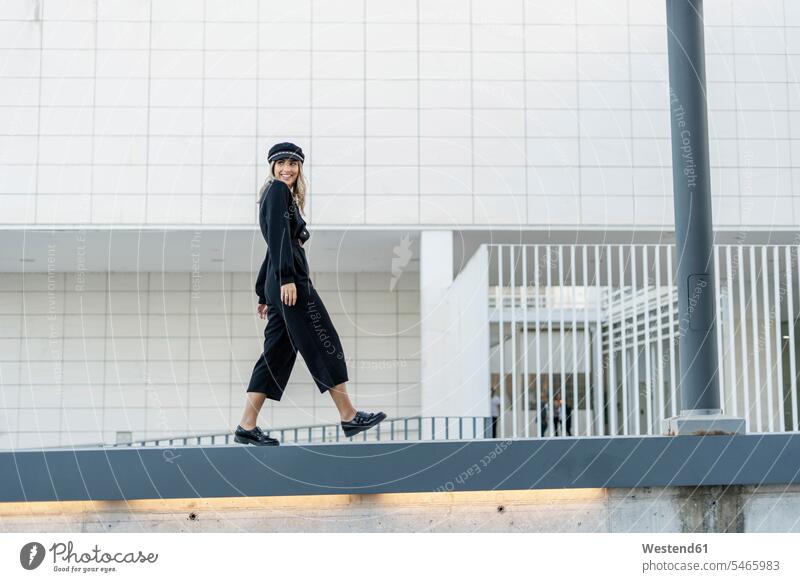 Junge blonde Geschäftsfrau mit schwarzer Matrosenmütze, die auf einer Mauer läuft geschäftlich Geschäftsleben Geschäftswelt Geschäftsperson Geschäftspersonen