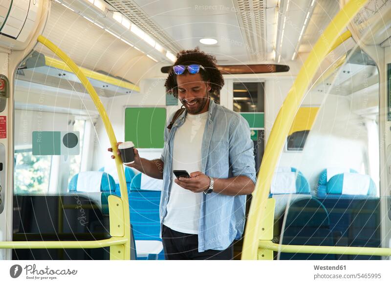 Junger trendbewusster Mann lächelt, während er im Zug ein Smartphone benutzt Farbaufnahme Farbe Farbfoto Farbphoto Fahrzeuginnenraum Zuginnenraum Bahn Züge