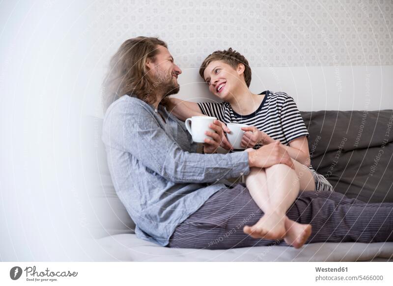 Glückliches junges Paar im Pyjama auf der Schlafcouch zu Hause Couch Couches Liege Sofas knuddeln schmusen sitzend sitzt früh Frühe Morgen entspannen relaxen
