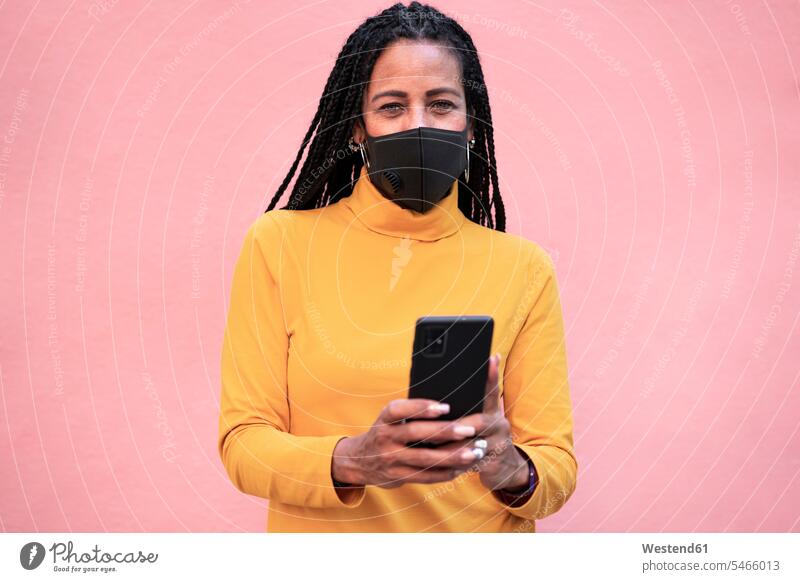 Reife Frau, die eine Gesichtsmaske trägt und ein Mobiltelefon benutzt, während sie an einer rosa Wand steht Farbaufnahme Farbe Farbfoto Farbphoto Außenaufnahme