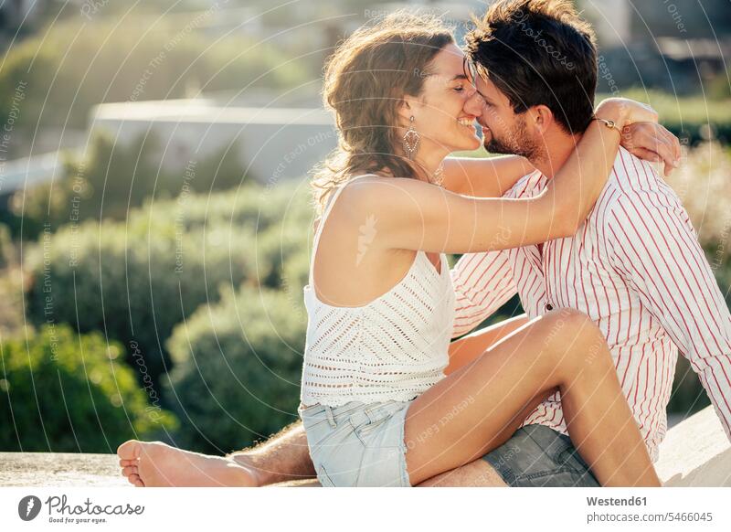 Romantisches Paar, das auf einer Mauer sitzt, sich umarmt und den Urlaub genießt Kuss Küsse sitzend Arm umlegen Umarmung Umarmungen Jahreszeiten sommerlich