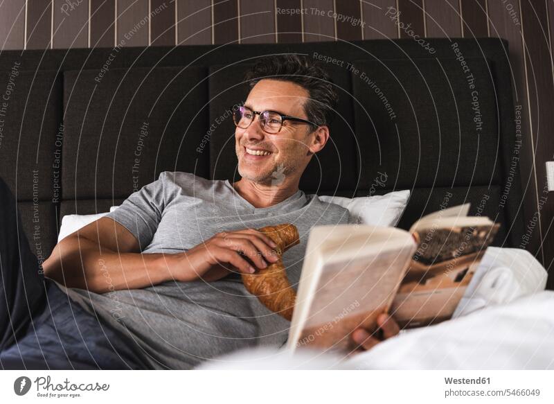 Glücklicher Mann liegt zu Hause im Bett mit Croissant und Buch glücklich glücklich sein glücklichsein Männer männlich Betten liegen liegend lesen Lektüre Bücher