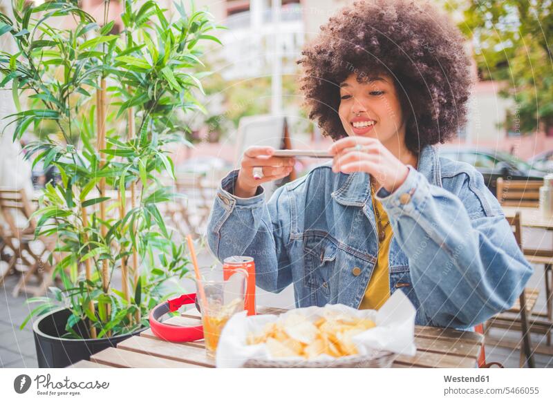 Lächelnde junge Frau mit Afrofrisur beim Fotografieren mit dem Smartphone in einem Straßencafé in der Stadt Leute Menschen People Person Personen gelockt