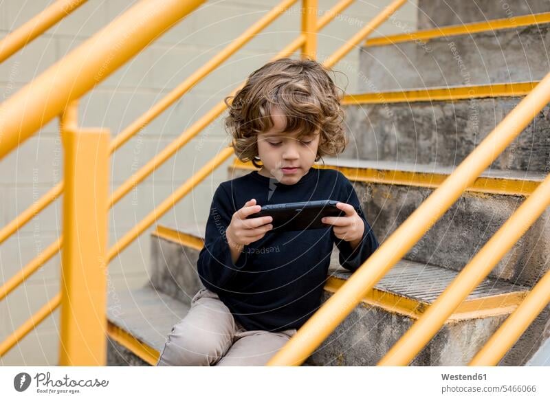 Junge sitzt auf der Treppe und spielt mit Handheld-Spielkonsole Handehelds sitzen sitzend Konsole spielen Treppenaufgang Buben Knabe Jungen Knaben männlich Kind