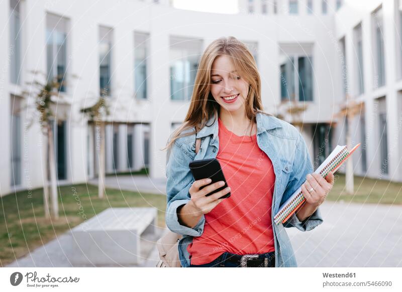 Lächelnde junge blonde Studentin benutzt Smartphone an der Universität schöne Frau schöne Frauen Mensch Menschen Leute People Personen Campus